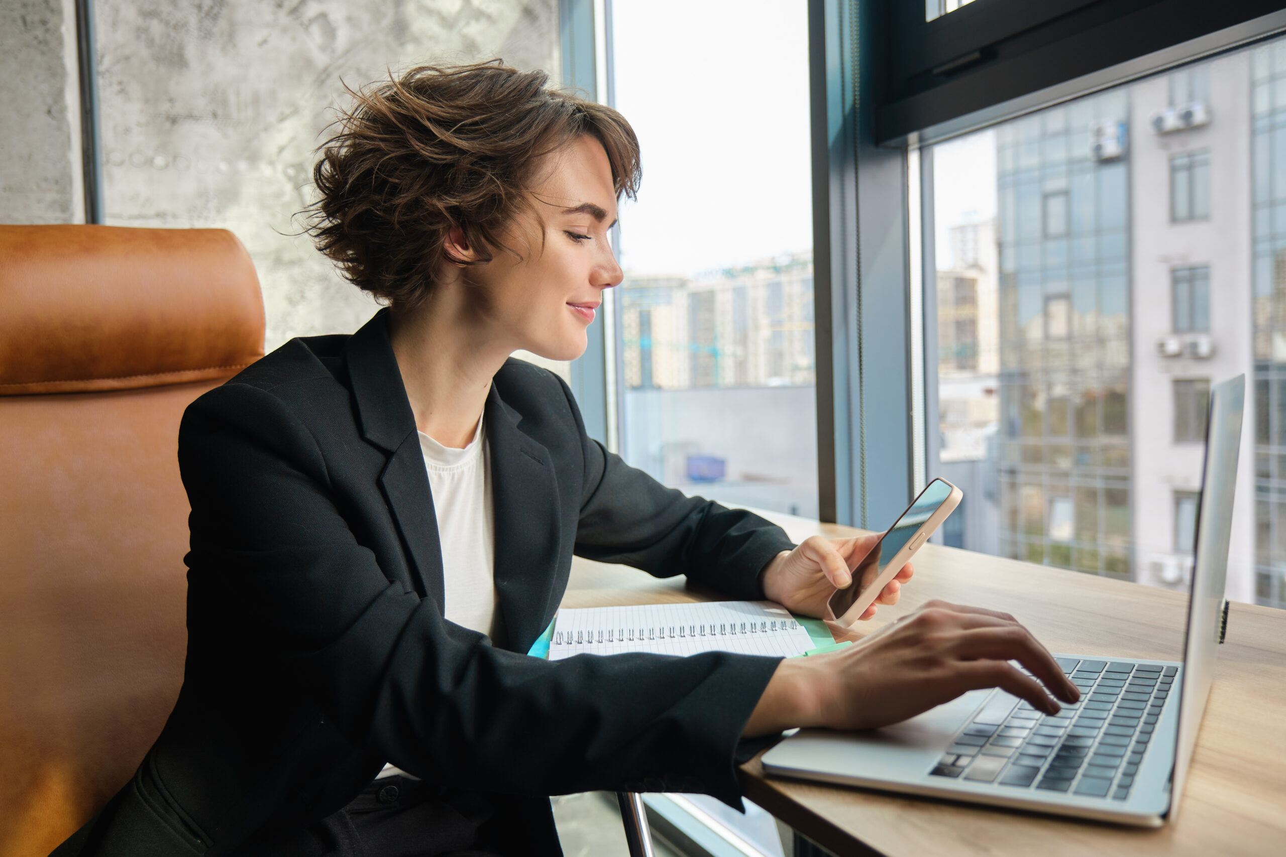 Young working woman in office, sitting in front of computer and using smartphone, wearing suit